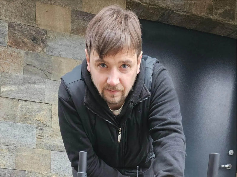 Man with brown hair and beard in black jacket leans forward, looking at camera, stone wall and dark door behind him.