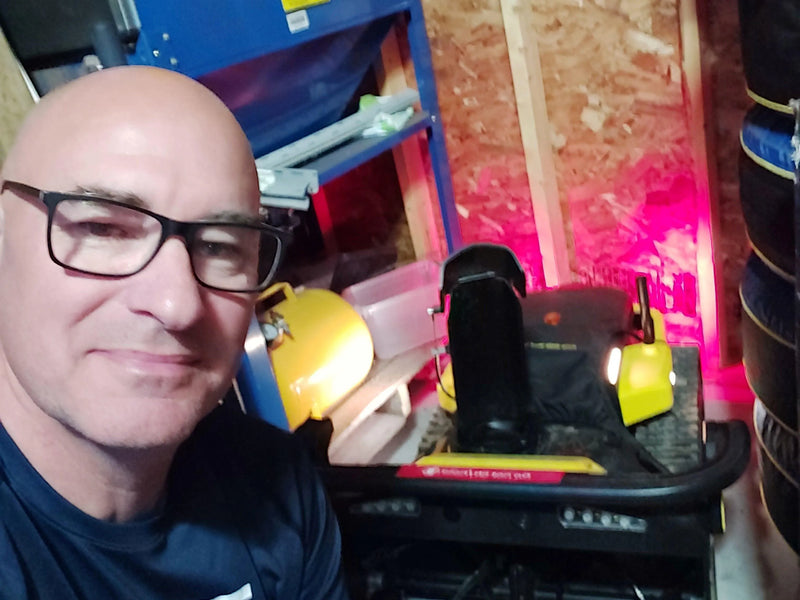 Bald man with glasses smiles in garage selfie, yellow lawn mower and blue shelf in background, pink light on wall.