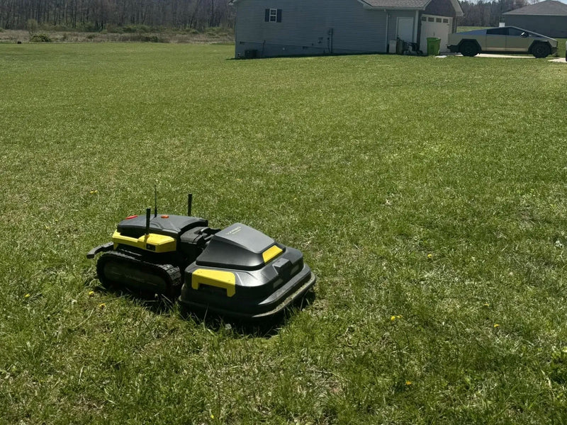 Two black and yellow robotic lawn mowers on a grassy yard, with a gray house and silver Tesla Cybertruck in the background.