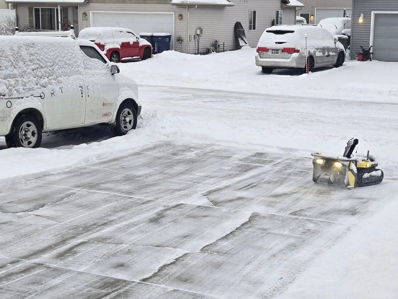 Snow-covered driveway with cleared concrete sections; yellow snow blower in foreground, white and red vehicles parked, houses in background.