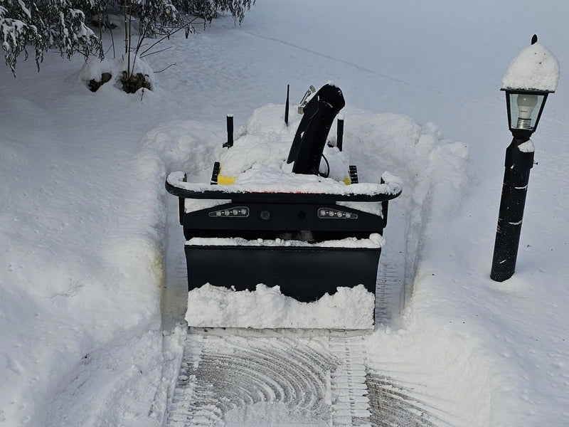 Black snow blower clearing path in deep snow, surrounded by snow-covered ground and lamp post.