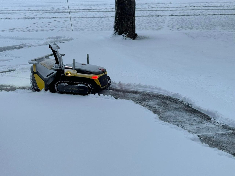 Yellow and black robotic lawn mower with illuminated lights clearing snow on a driveway, tree in background.