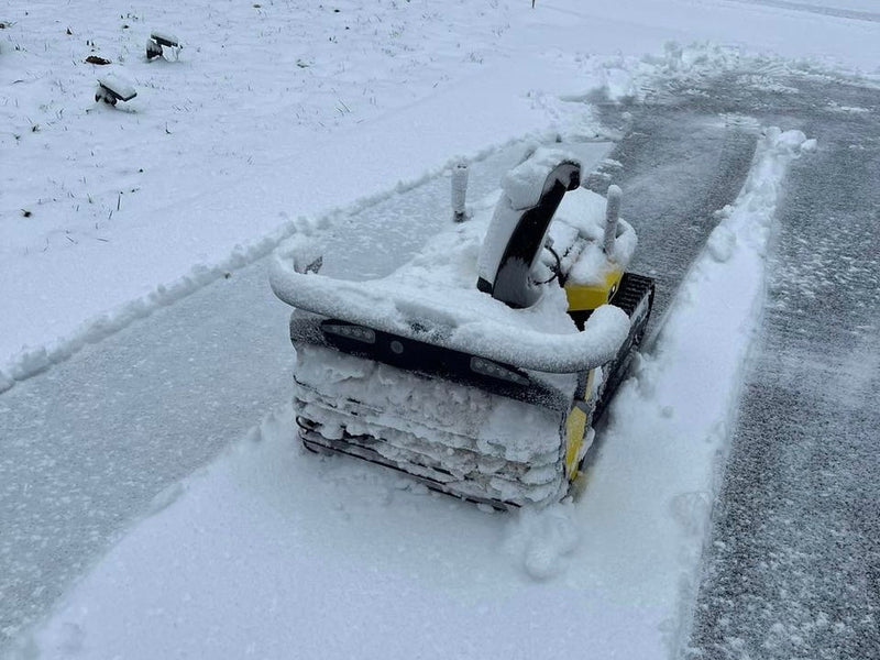 Snow-covered yellow and black snowplow clearing path on icy road, surrounded by deep snow.