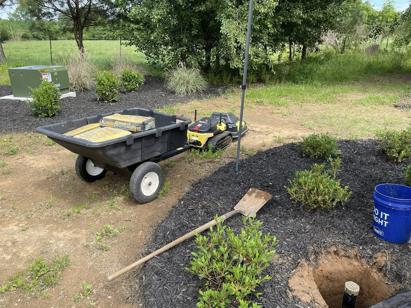 Black wheelbarrow with Quikrete bags, yellow robotic mower, shovel, blue Lowe's bucket, and mulched garden beds in outdoor yard.