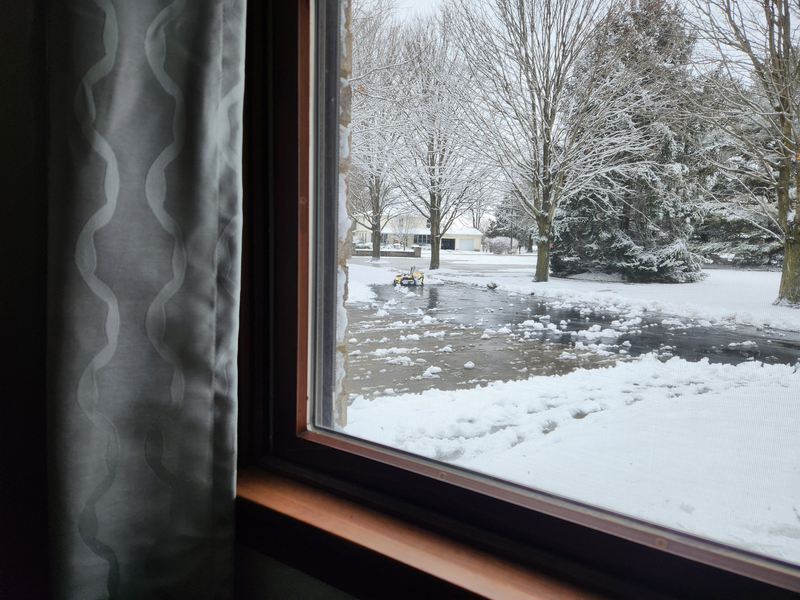 Gray curtain beside window showing snowy yard with bare trees, snow-covered ground, and yellow snowblower in distance.