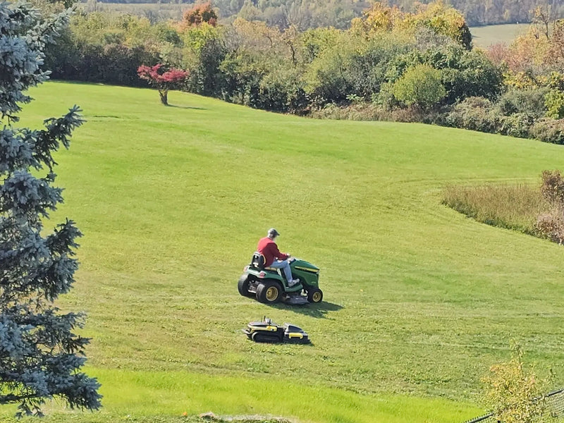 Man in red shirt mowing large green lawn with riding mower, robotic mower nearby, trees in background.