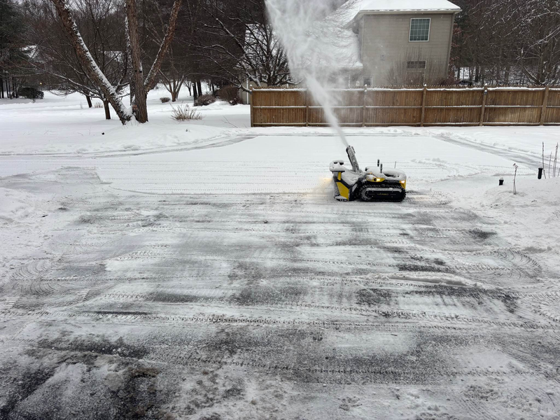 Yellow and black snow blower clearing driveway, ejecting snow to right, with snow-covered trees and house in background.