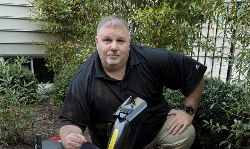 Man in black polo shirt kneels outdoors holding black equipment, surrounded by green shrubs and house siding.