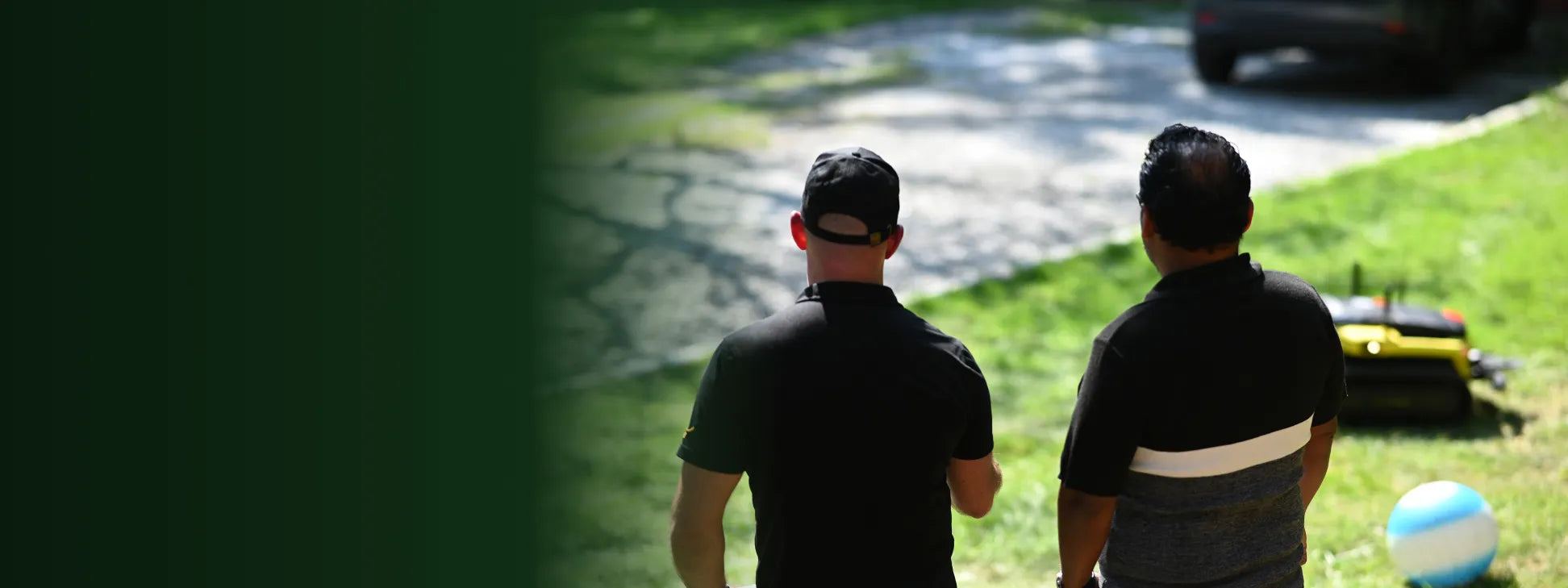 Two men in black shirts walk away on a sunny day, one wearing a cap, near a yellow lawnmower and blue-white ball on grass.
